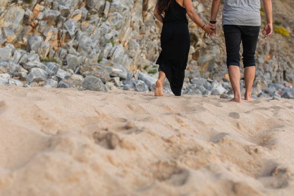 cropped view of couple holding hands and walking on sand near rocks  - Photo, Image