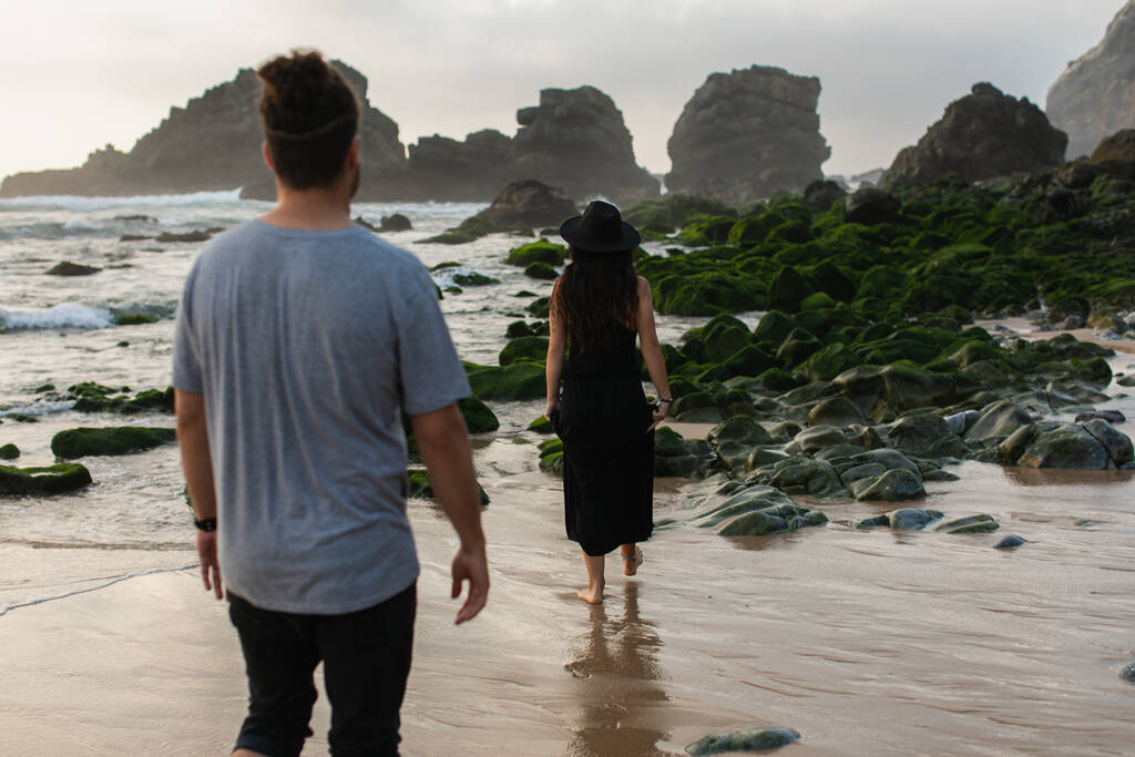 visão traseira do homem andando atrás de mulher tatuada em vestido preto perto de musgo verde em pedras e oceano  - Foto, Imagem