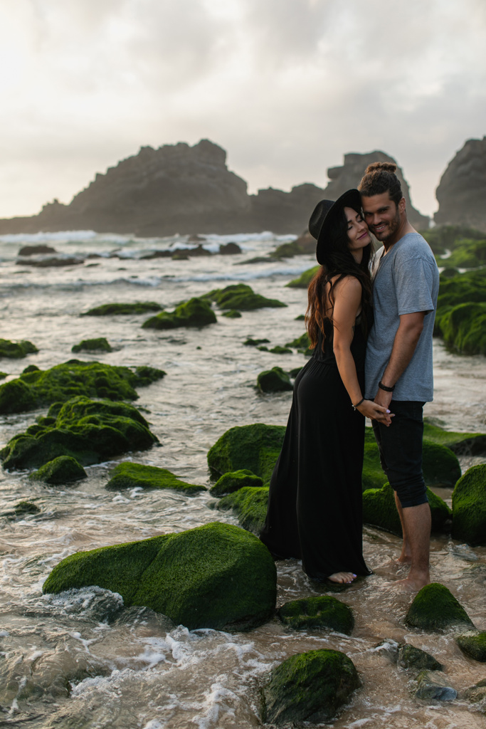 pleased woman in hat and dress holding hands with bearded boyfriend near mossy stones and ocean - Photo, Image