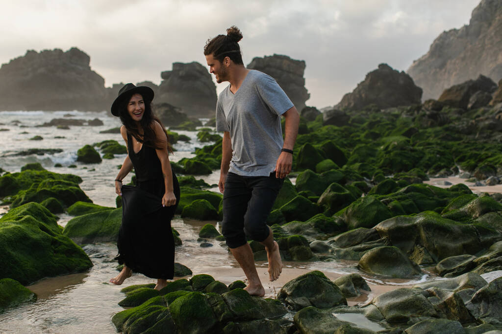 szakállas férfi és vidám nő ruha és kalap séta közelében mohás kövek óceán - Fotó, kép