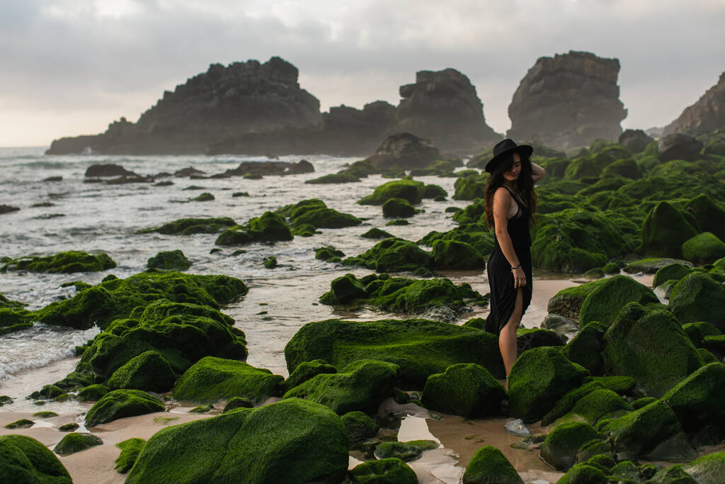 vista panorámica de la mujer tatuada en vestido y sombrero de pie cerca de las piedras verdes musgosas en el océano - Foto, Imagen