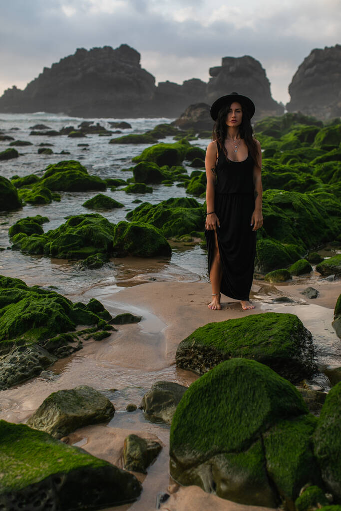 mujer tatuada en vestido negro y sombrero de pie cerca de piedras verdes musgosas en el océano - Foto, Imagen