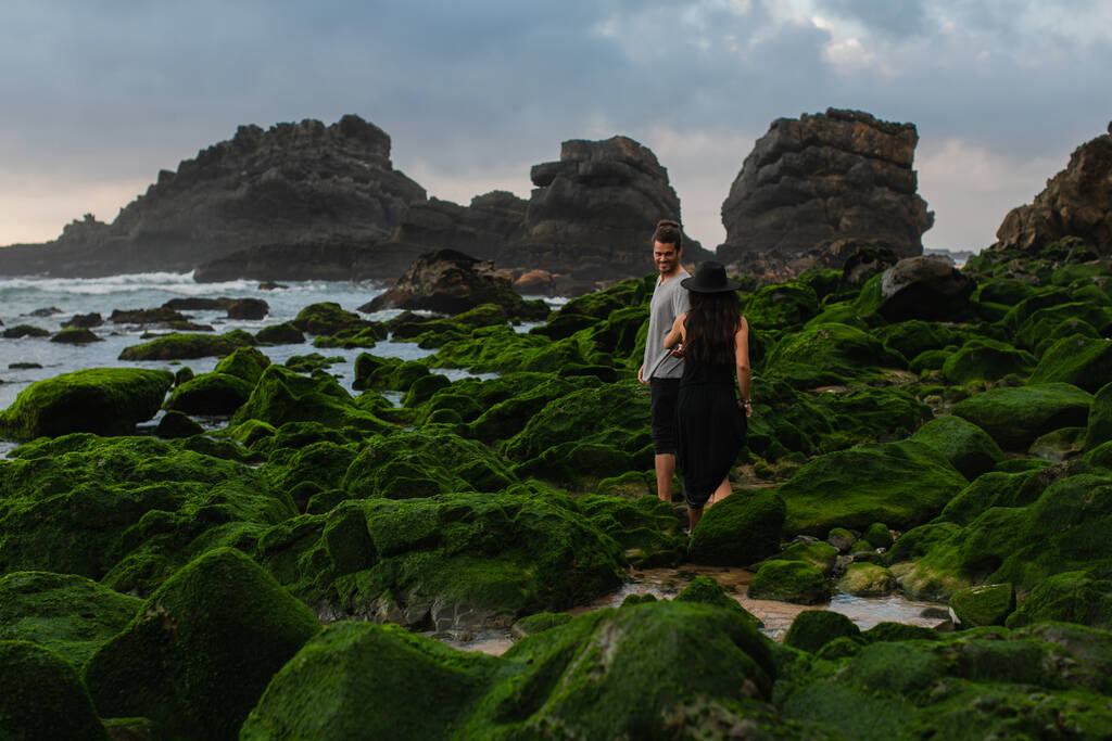 Şapkalı ve elbiseli bir kadının, okyanus kıyısındaki yosunlu taşların üzerinde mutlu bir adamın yanında yürüdüğünü. - Fotoğraf, Görsel