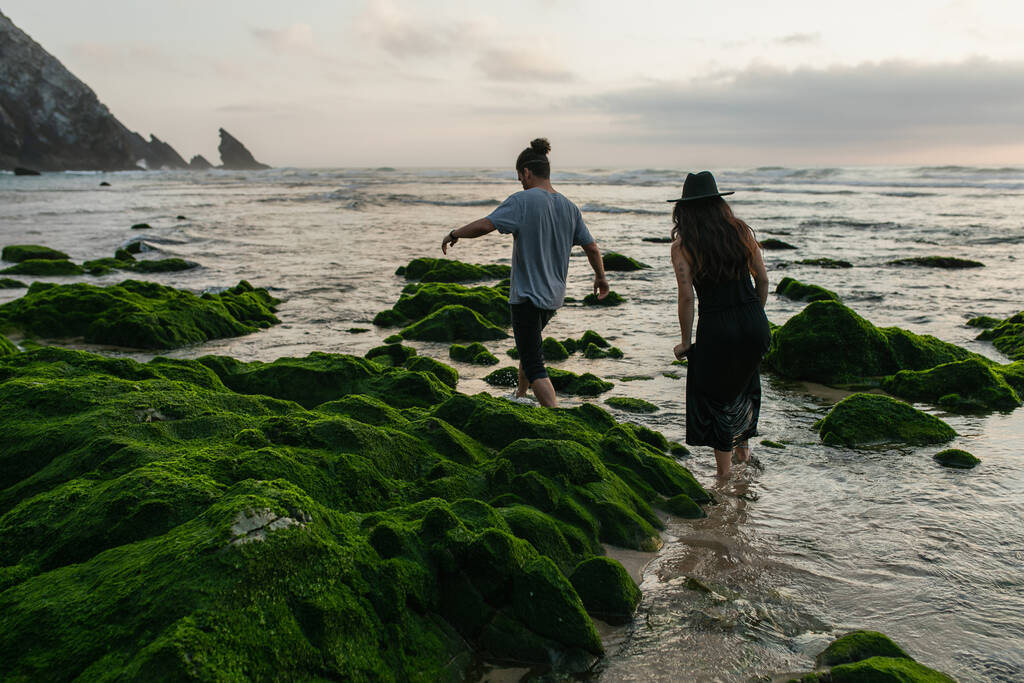 вид ззаду пари, що йде біля зелених мохоподібних каменів в океані
 - Фото, зображення