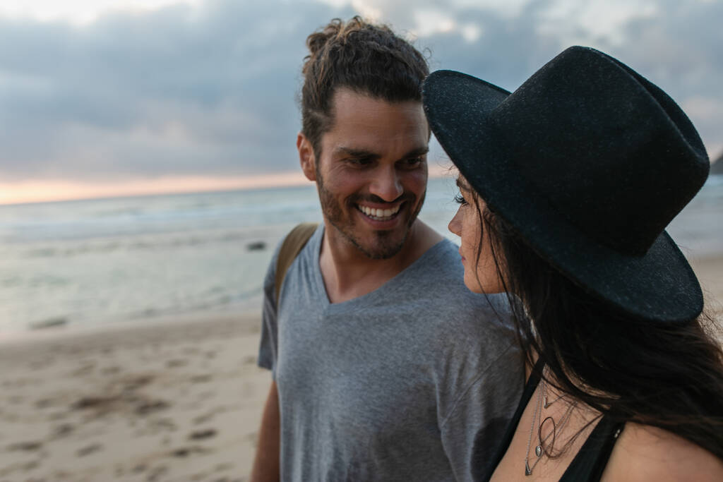 vidám szakállas férfi nézi nő fekete kalap a strandon  - Fotó, kép