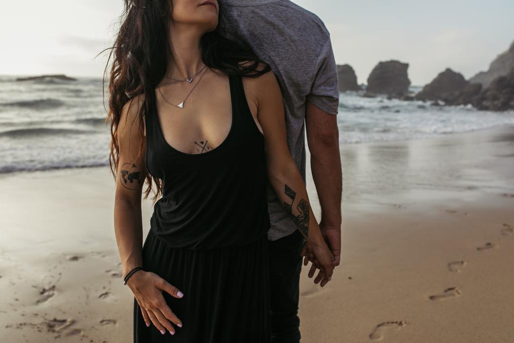 przycięte widok z tatuażem kobieta i mężczyzna trzymając się za ręce w pobliżu oceanu wieczorem - Zdjęcie, obraz