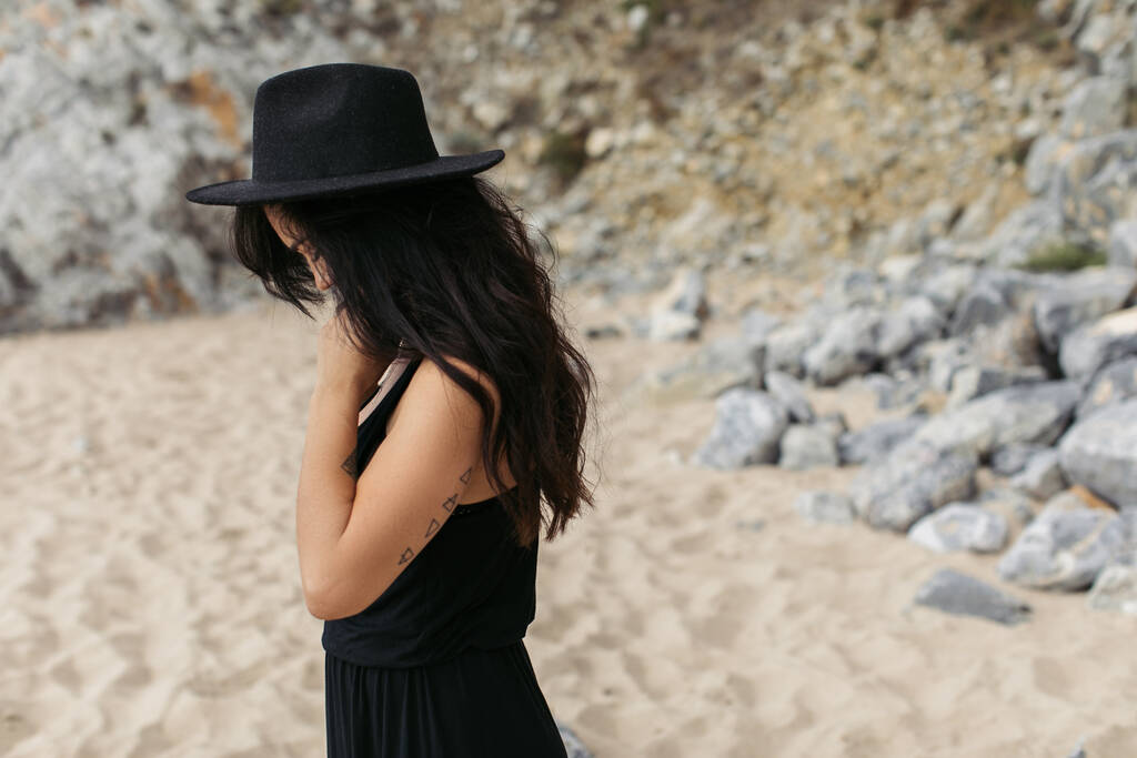 tetovált nő ruha és fekete kalap áll homokos strandon Portugáliában  - Fotó, kép