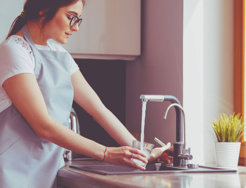 Menschliche Hand hält Glas und gießt frisches Trinkwasser an den Küchenhahn. - Foto, Bild
