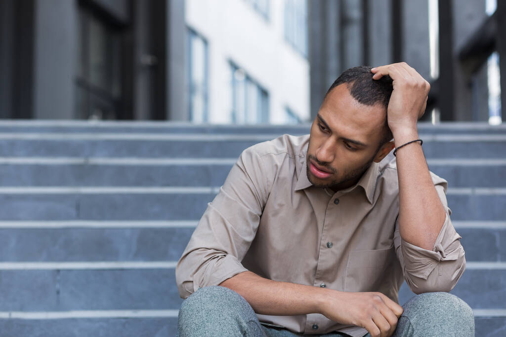 Disappointed and sad man sitting on stairs outside office building, employee fired from work, african american man in shirt upset and depressed. - Photo, image