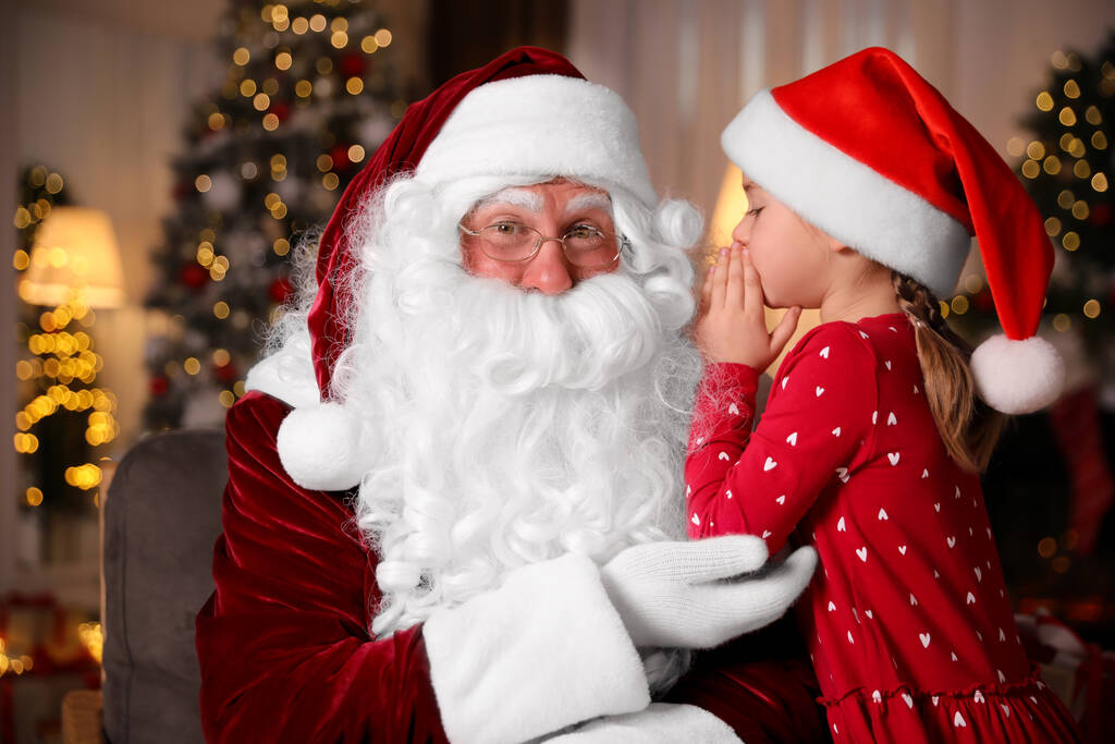 Маленькая девочка шепчет в ухо Санта Клаусу возле рождественской елки в помещении - Фото, изображение