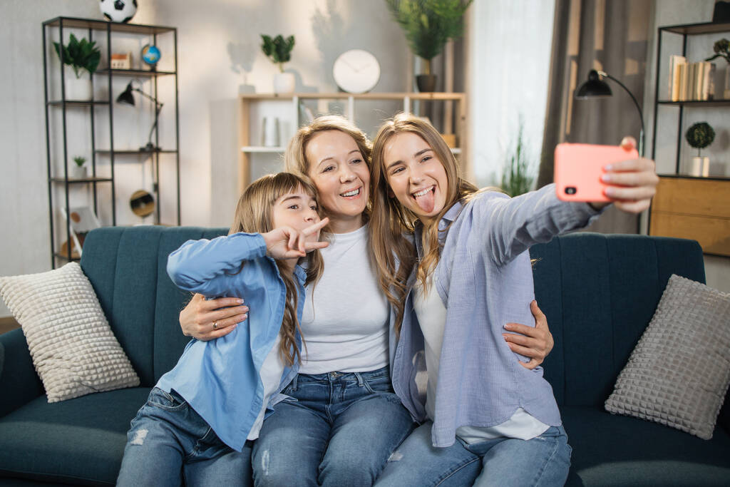 Head shot portrait close up smiling two daughters with mother taking selfie using smart phone, showing their tongues, happy mom with adorable kids making video call or recording vlog - 写真・画像