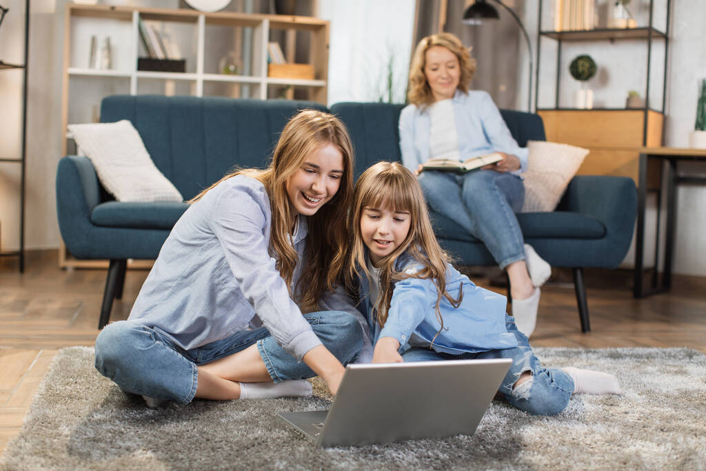 Cute blond girls sisters sitting on warm floor play at home together using laptop, while happy mother relax on sofa on background. - Foto, imagen