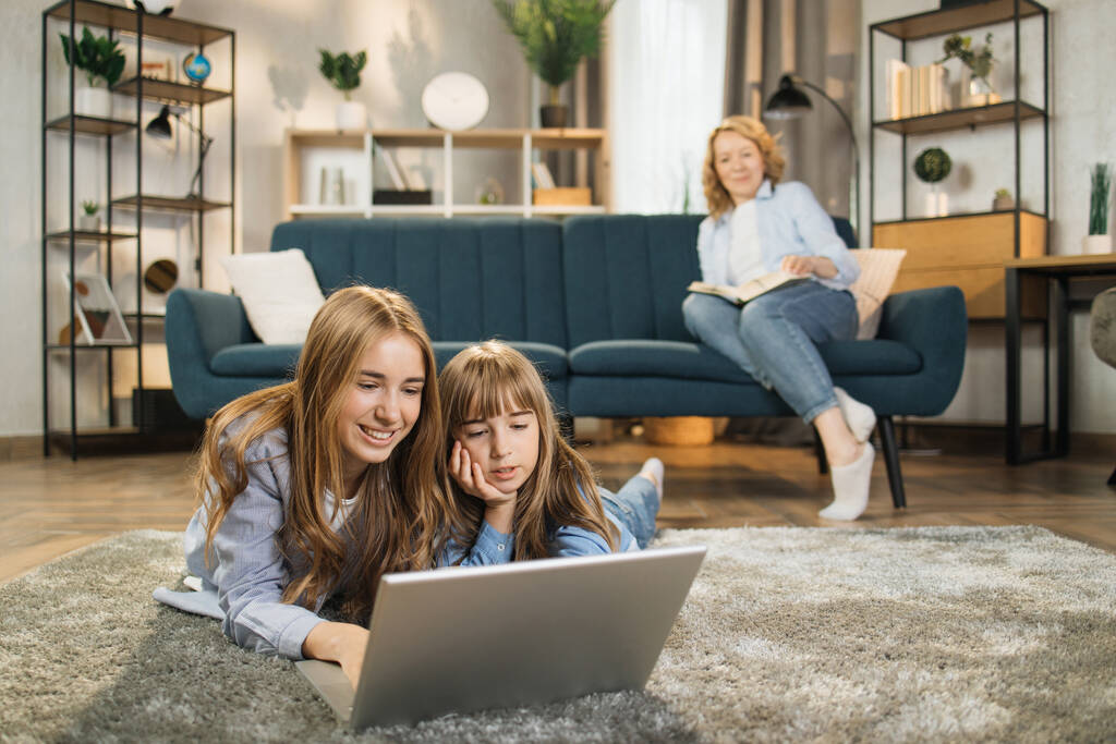 Happy family leisure at home concept, young mother relaxing reading book on sofa in comfort living room while cheerful kids daughters enjoying free time to watch movie lying on warm floor - Foto, imagen