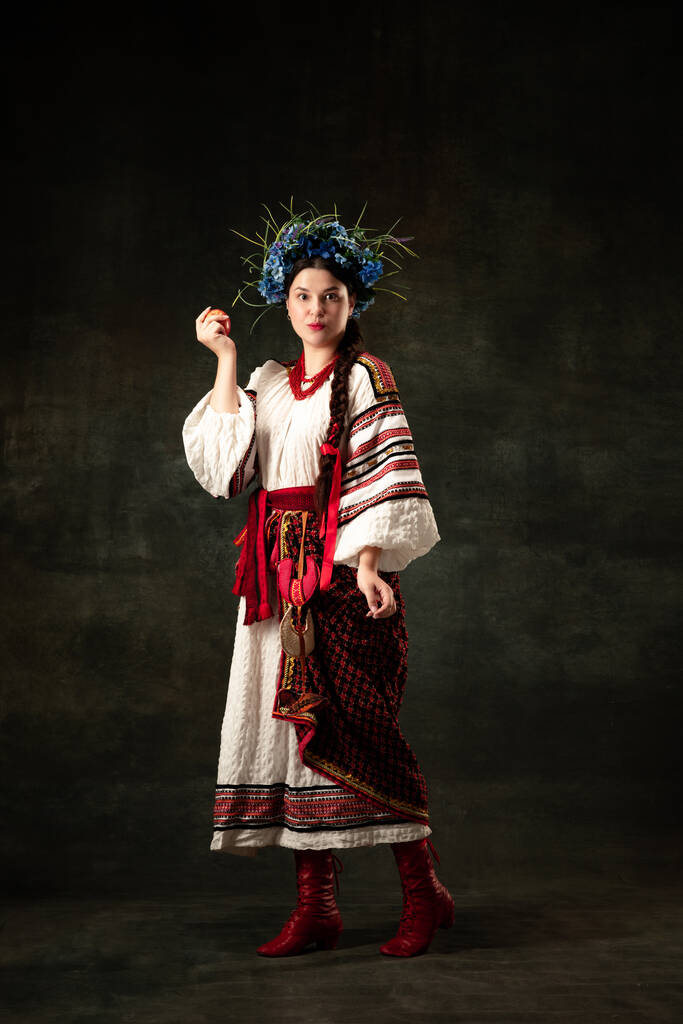 Emotional adorable woman wearing national folk Ukrainian attire posing isolated over dark vintage background. Fashion, beauty, cultural heritage concept. - Fotoğraf, Görsel