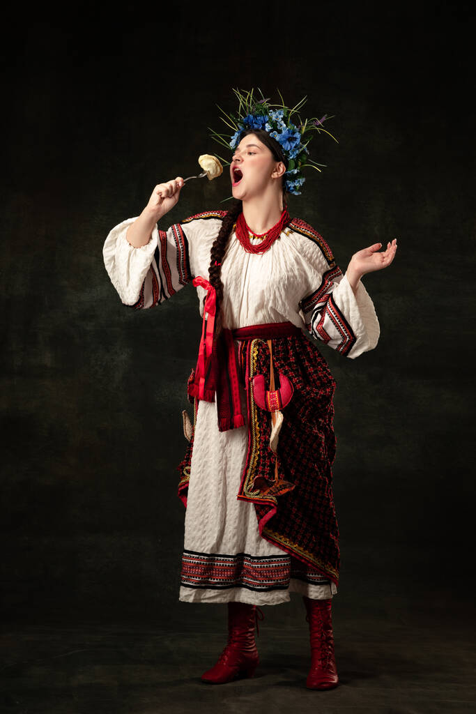 Traditions, national cuisine. Creative portrait of beautiful Ukrainian woman in traditional folk costume tasting dumpling isolated over dark background. Fashion, beauty, cultural heritage concept. - Valokuva, kuva