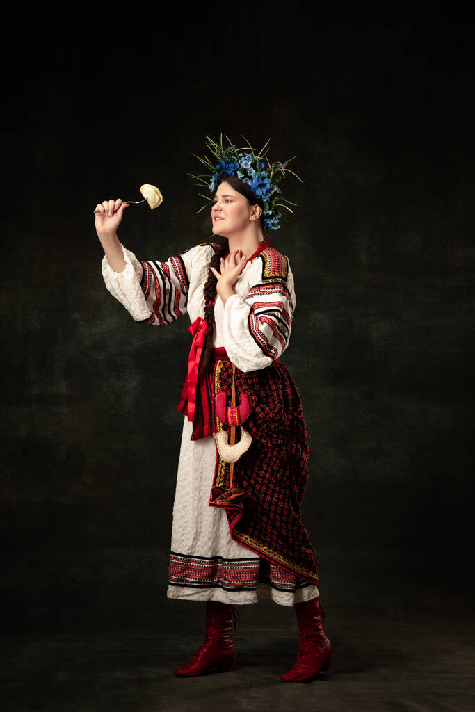 Traditions, national cuisine. Creative portrait of beautiful Ukrainian woman in traditional folk costume tasting dumpling isolated over dark background. Fashion, beauty, cultural heritage concept. - Фото, зображення