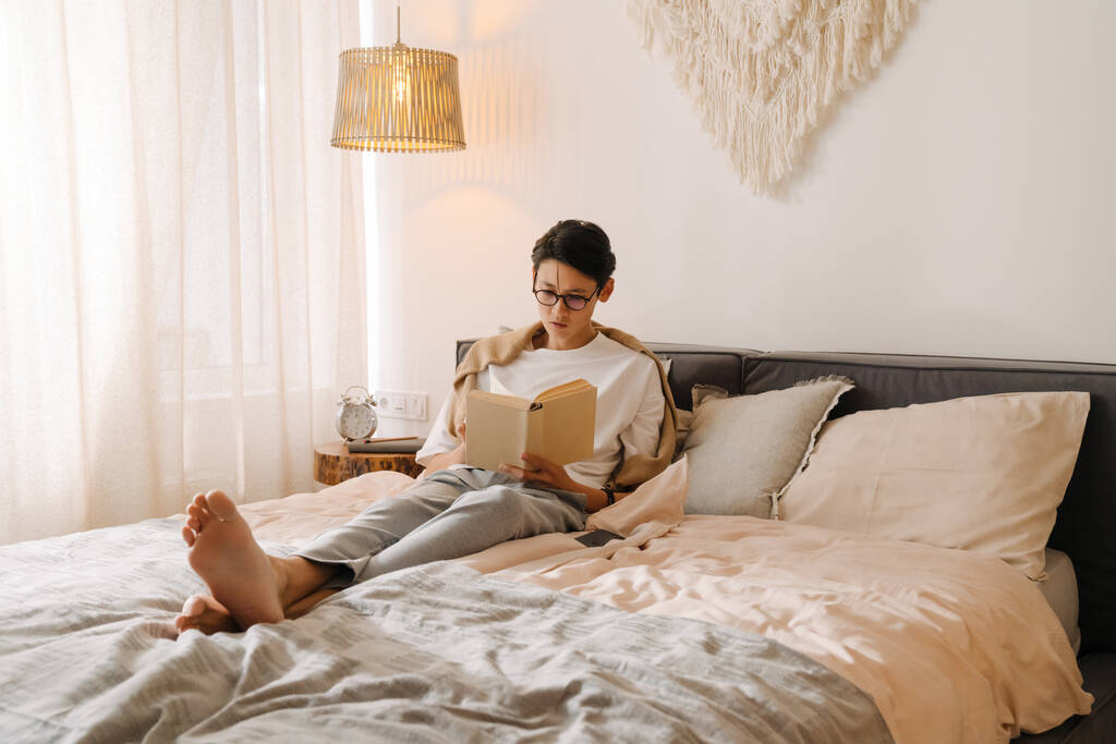 Asian boy in eyeglasses reading book while resting on bed at home - Фото, изображение