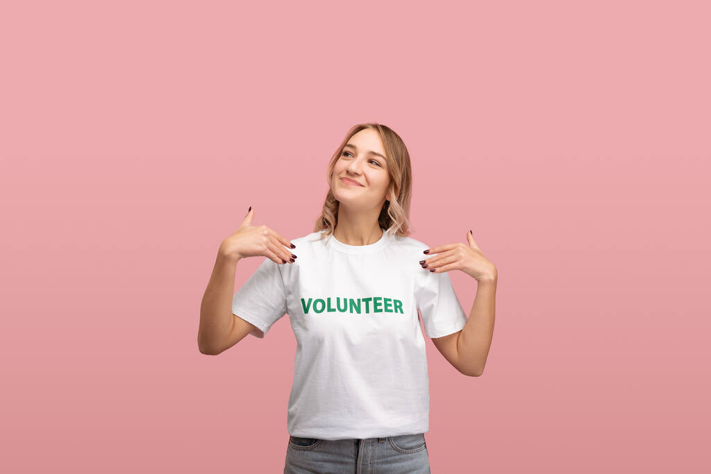 Iloinen nuori nainen katselee pois hymyillen ja osoittaa vapaaehtoisen kirjoitus valkoisella t-paidalla vaaleanpunaista taustaa vasten - Valokuva, kuva