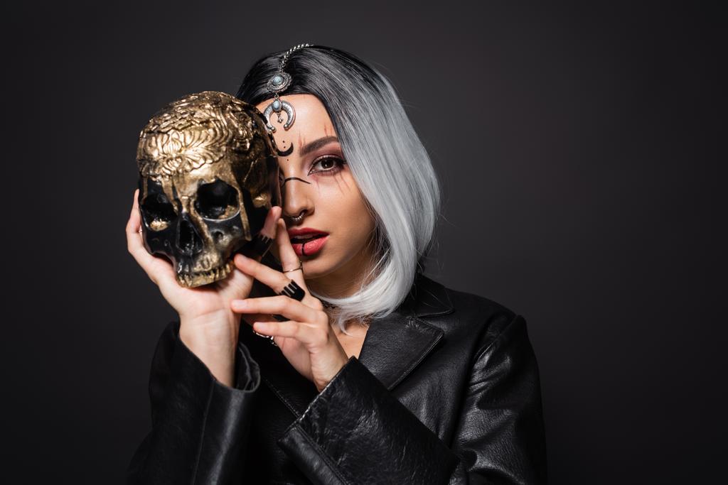 молодая женщина в стиле ведьмы с золой светлые волосы затмевают лицо с жутким золотистым черепом изолированы на черном - Фото, изображение