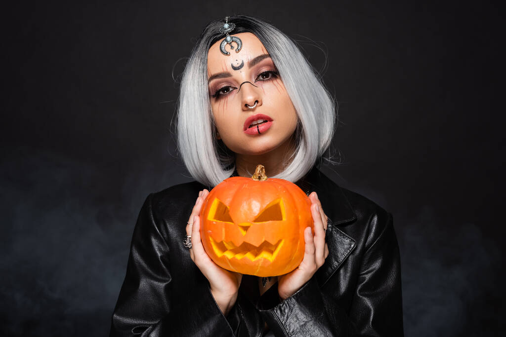 young ash blonde woman with witch makeup holding jack o lantern on black background - Photo, Image