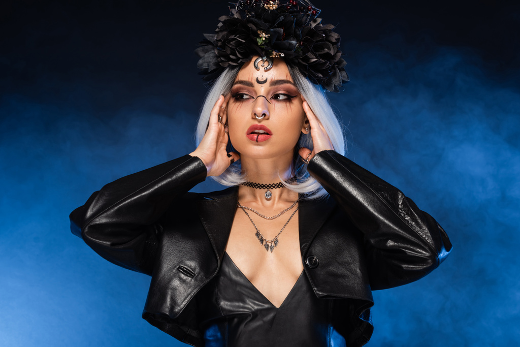 mujer sexy en corona negra y maquillaje de bruja tocando ceniza pelo rubio sobre fondo oscuro con humo azul - Foto, Imagen