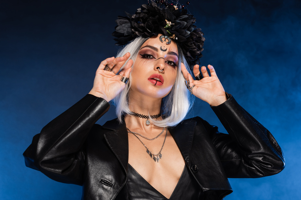 sexy mujer en bruja halloween traje y corona tocando ceniza pelo rubio sobre fondo azul oscuro - Foto, imagen