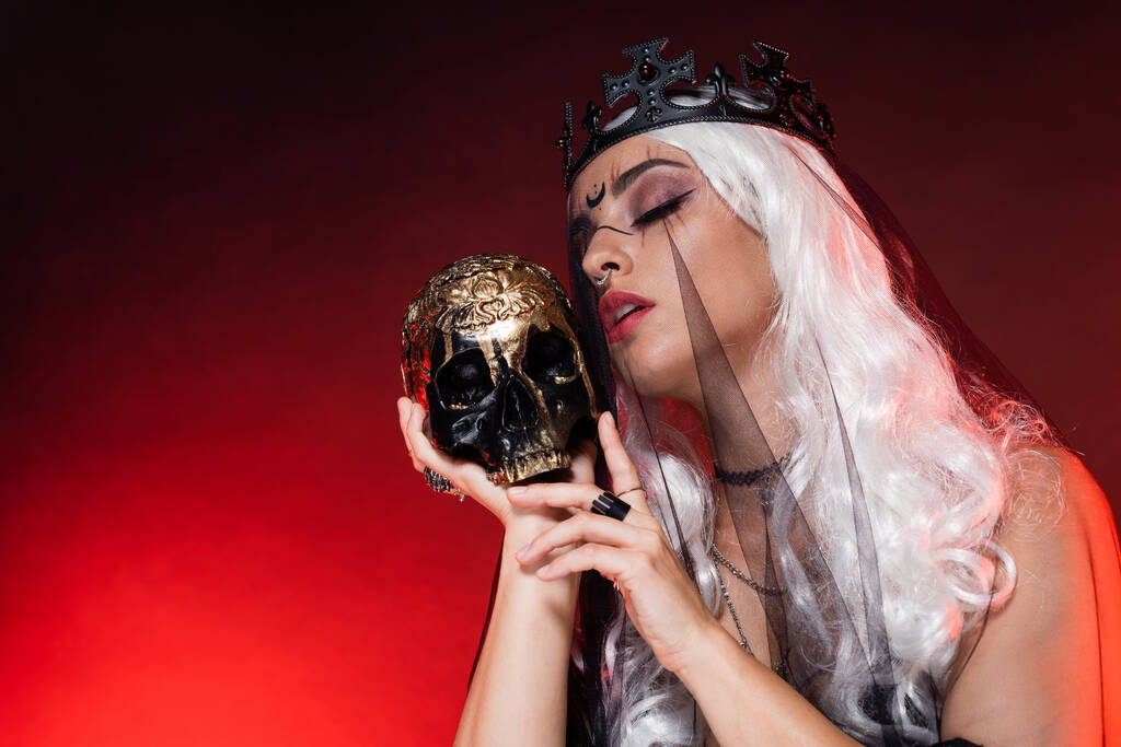 blonde woman with closed eyes in witch halloween makeup and crown near golden skull on dark red background - Photo, Image