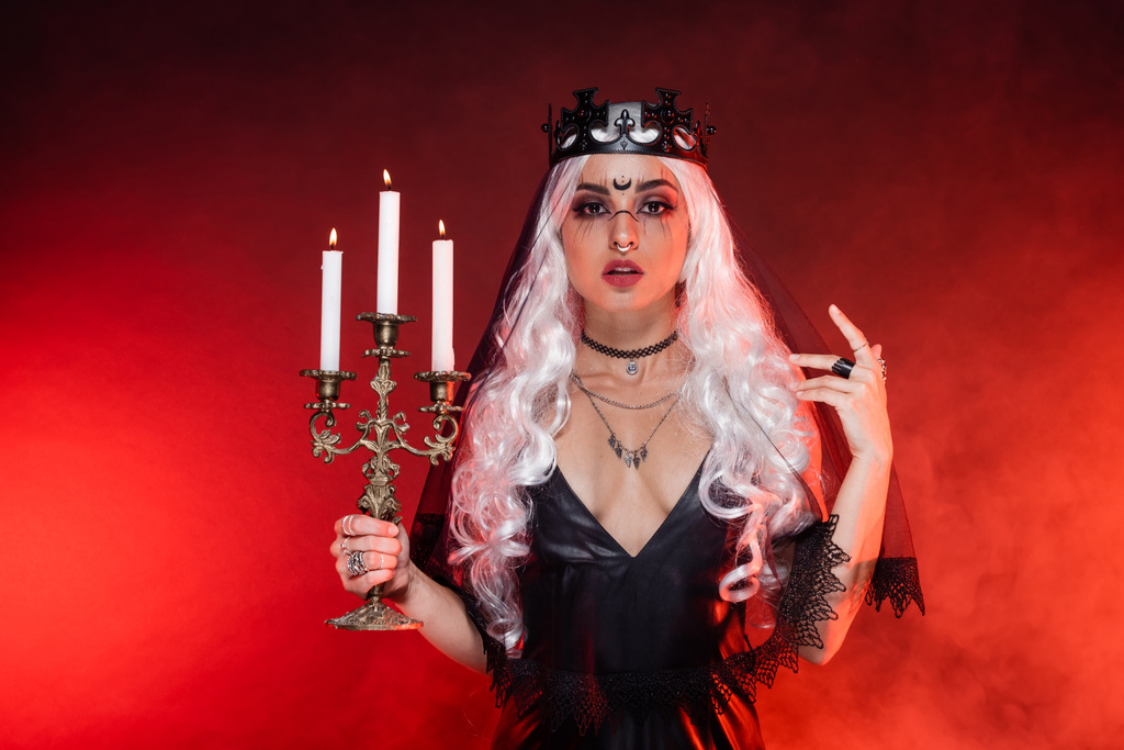 blonde woman in black dress and witch crown holding triple candlestick on background with red smoke - Photo, Image