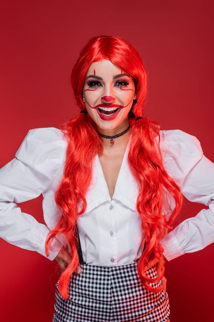 usmívající se žena s klaunem make-up a světlé vlasy stojící s rukama na bocích izolovaných na červené - Fotografie, Obrázek