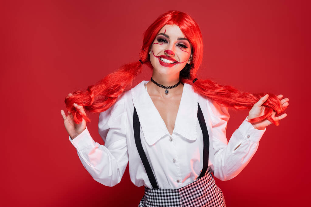 весела жінка з клоуном Хеллоуїн макіяж тримає яскраві хвостики ізольовані на червоному
 - Фото, зображення