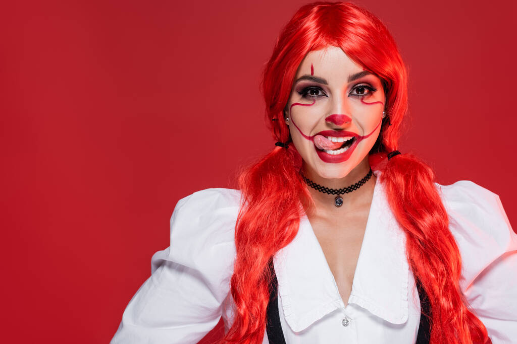 mujer alegre con pelo largo brillante y maquillaje payaso sobresaliendo lengua aislada en rojo - Foto, imagen
