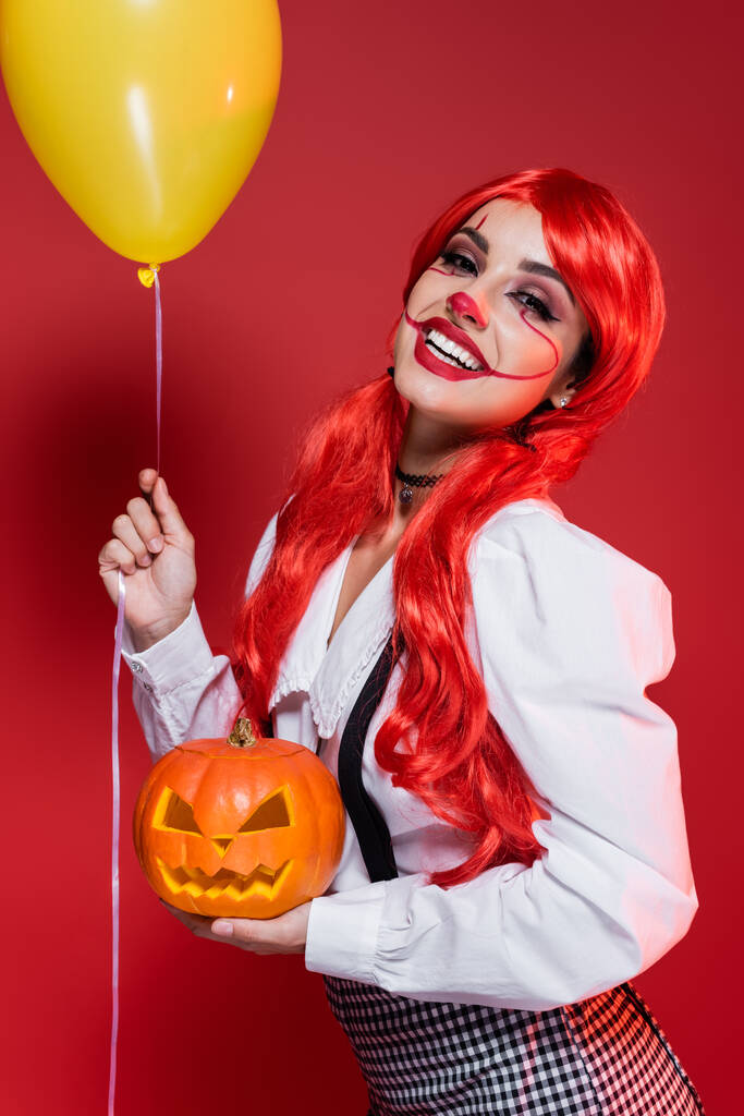 χαρούμενη γυναίκα με φωτεινά μαλλιά και αποκριάτικο μακιγιάζ κρατώντας κίτρινο μπαλόνι και σκαλιστά κολοκύθα απομονώνονται σε κόκκινο - Φωτογραφία, εικόνα