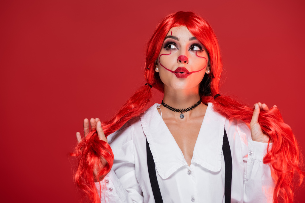 ošemetná žena s klaunem make-up drží světlé culíky a hledá izolované na červené - Fotografie, Obrázek