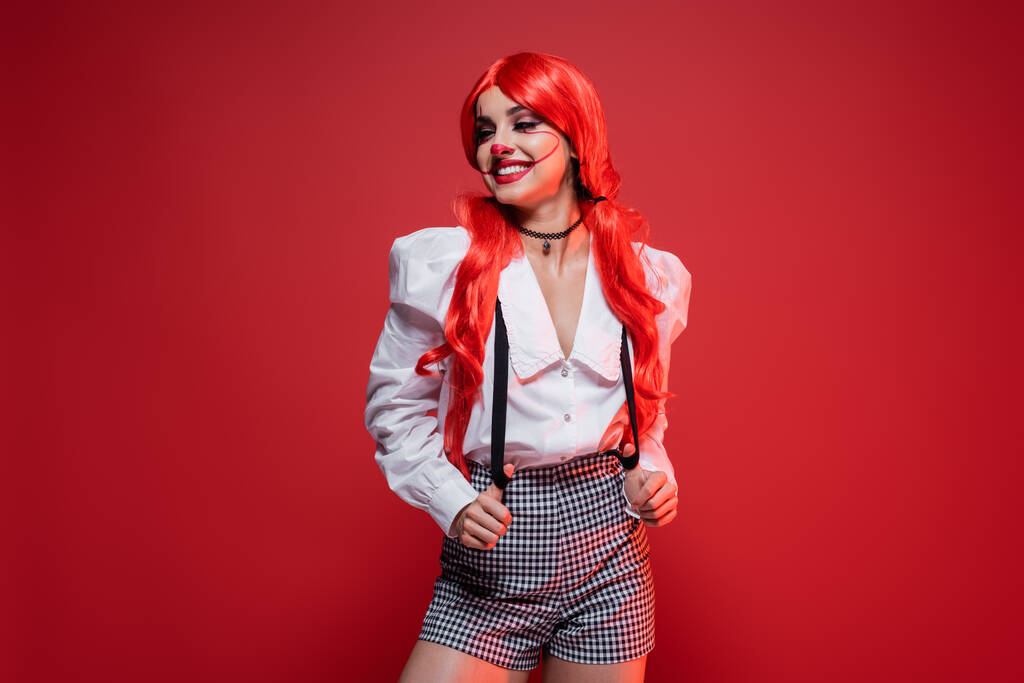 redhead woman with clown makeup posing in white blouse and shorts with suspenders isolated on red - Photo, Image