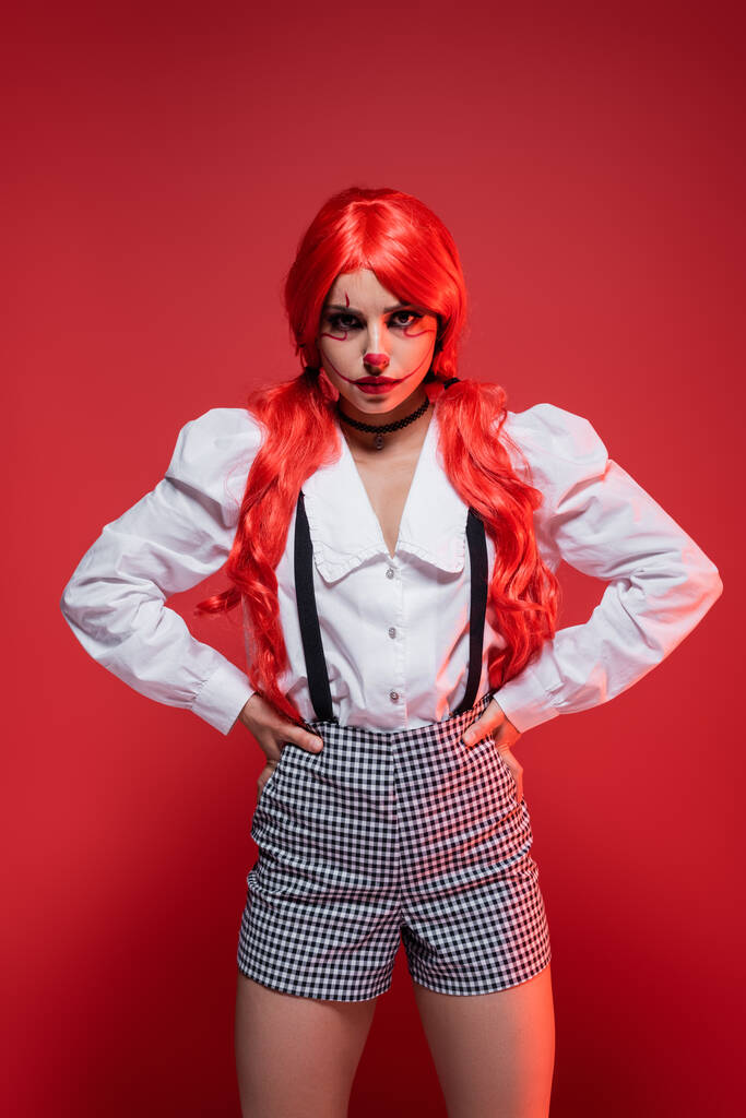 femme en colère avec des cheveux longs brillants et maquillage de clown debout avec les mains sur les hanches isolées sur rouge - Photo, image