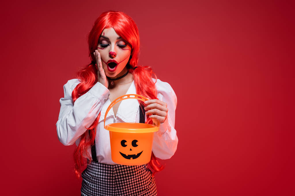 amazed woman with bright hair and clown makeup looking in halloween bucket isolated on red - Photo, Image