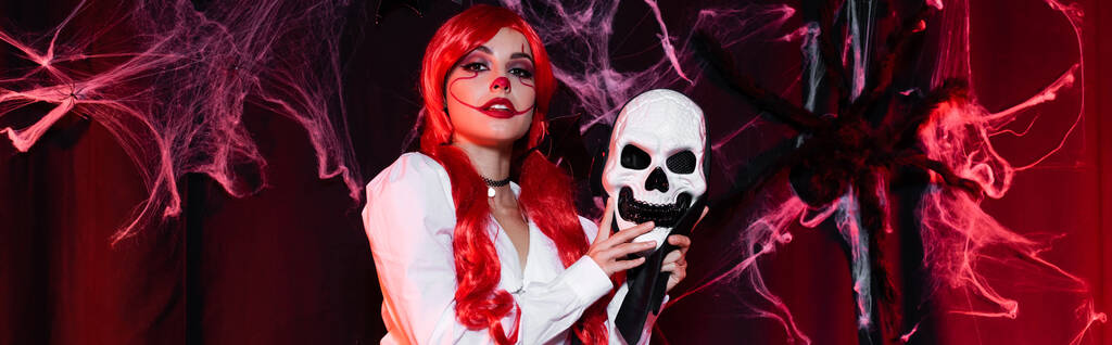рыжая женщина в клоунском макияже держа маску черепа возле паутины на темном фоне, баннер - Фото, изображение