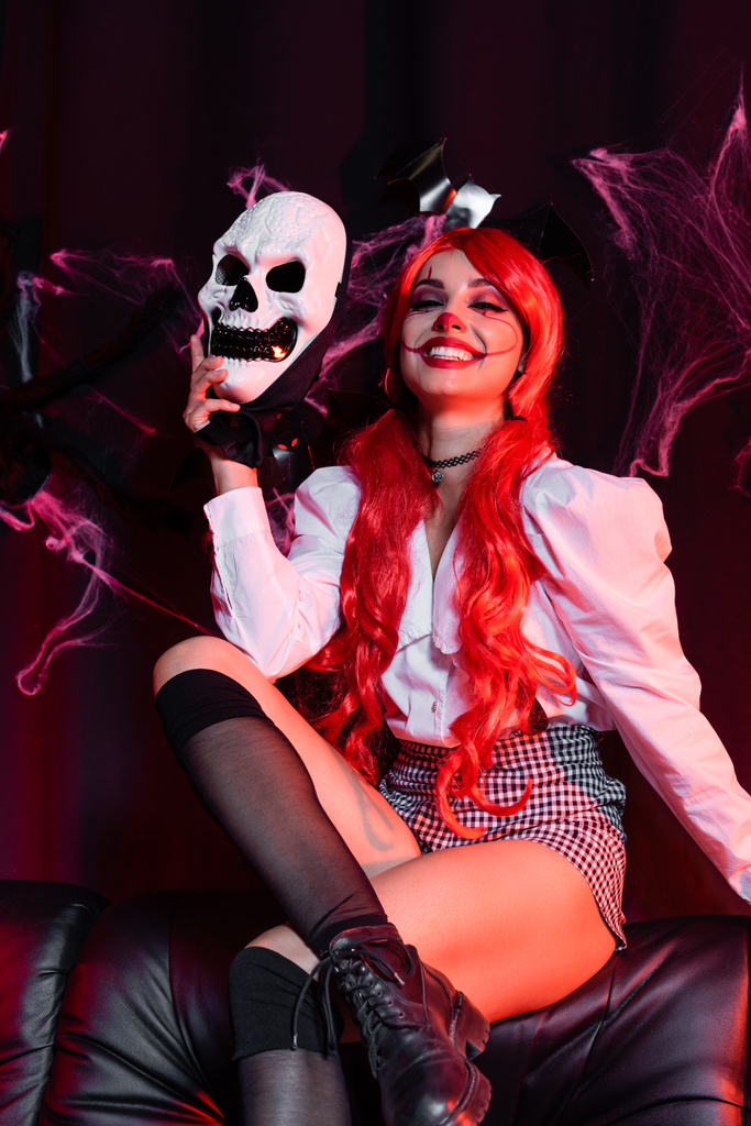 vrolijk roodharige vrouw in clown make-up houden schedel masker op zwarte achtergrond met spinnenweb - Foto, afbeelding
