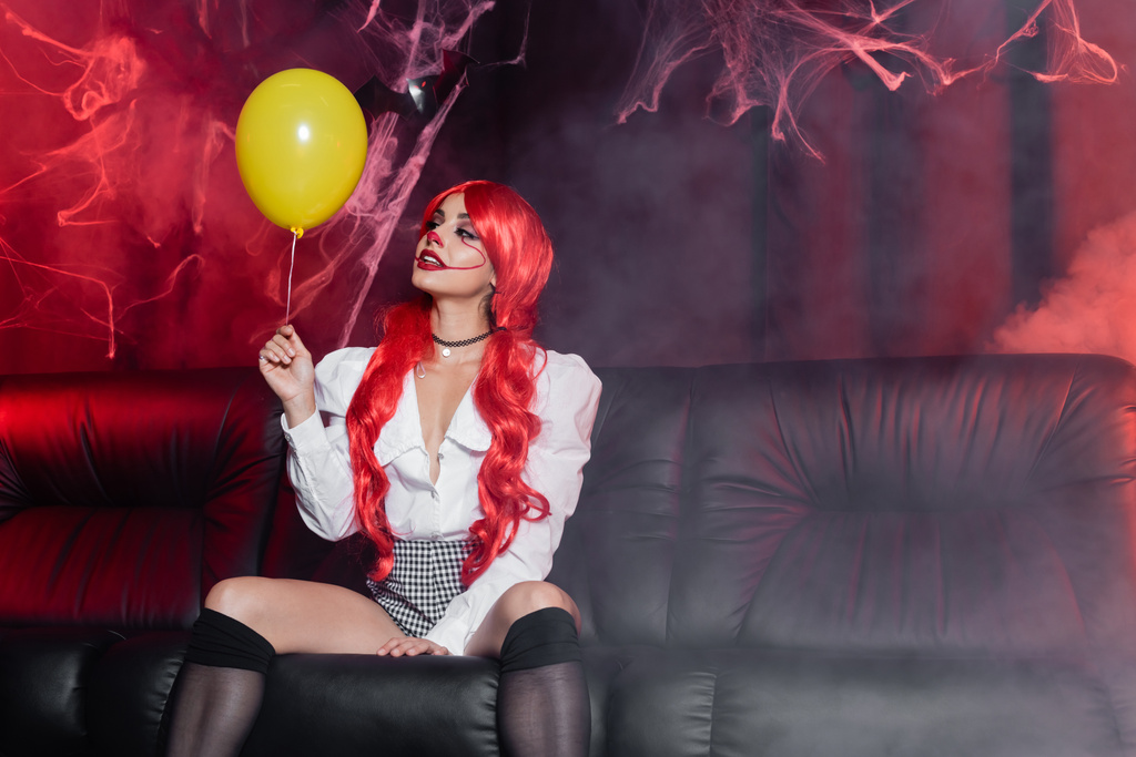 young redhead woman in clown makeup looking at yellow balloon on dark background with fog and spiderweb - Photo, Image