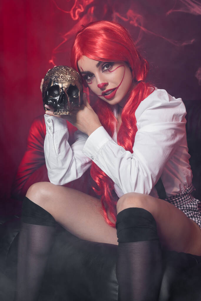 Mulher sexy com cabelo vermelho e maquiagem de palhaço olhando para a câmera perto do crânio dourado no fundo escuro com teia de aranha - Foto, Imagem