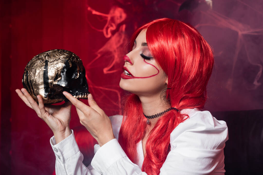 woman with red hair and clown makeup holding golden skull near spiderweb on dark red background - Photo, Image