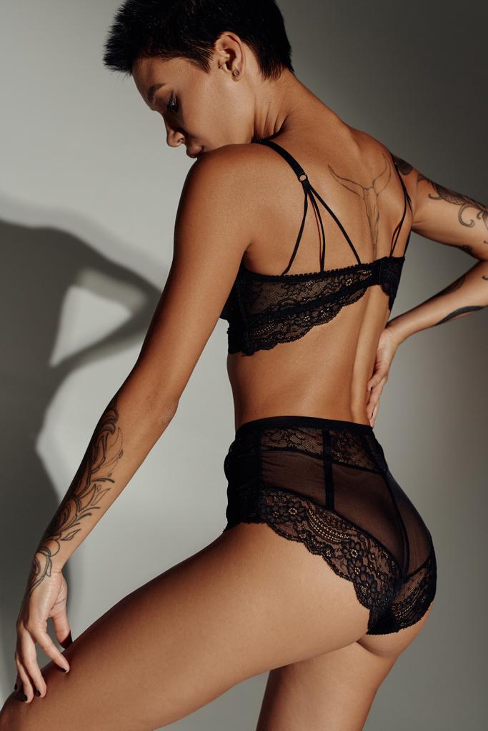 стройная татуированная женщина позирует в черном сексуальном белье на сером фоне с тенью - Фото, изображение