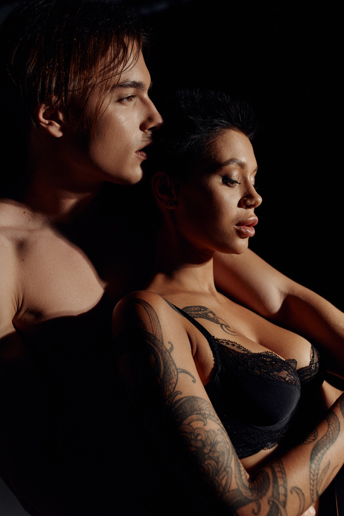 vista lateral de mujer tatuada con pecho sexy cerca de hombre sin camisa aislado en negro - Foto, Imagen