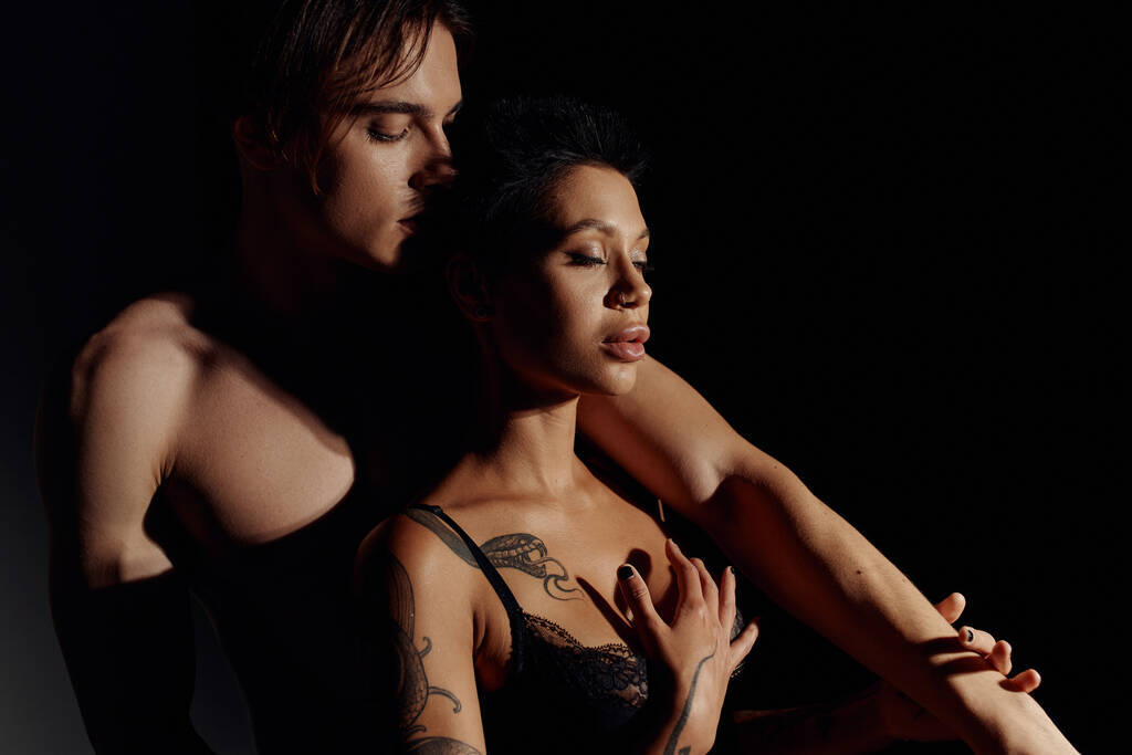 joven hombre sin camisa cerca sexy mujer tatuada en sujetador aislado en negro - Foto, imagen
