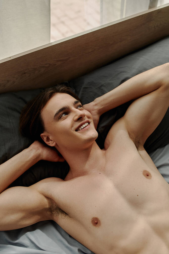 vista superior do jovem sexy olhando para longe e sorrindo enquanto deitado na cama com as mãos atrás da cabeça - Foto, Imagem