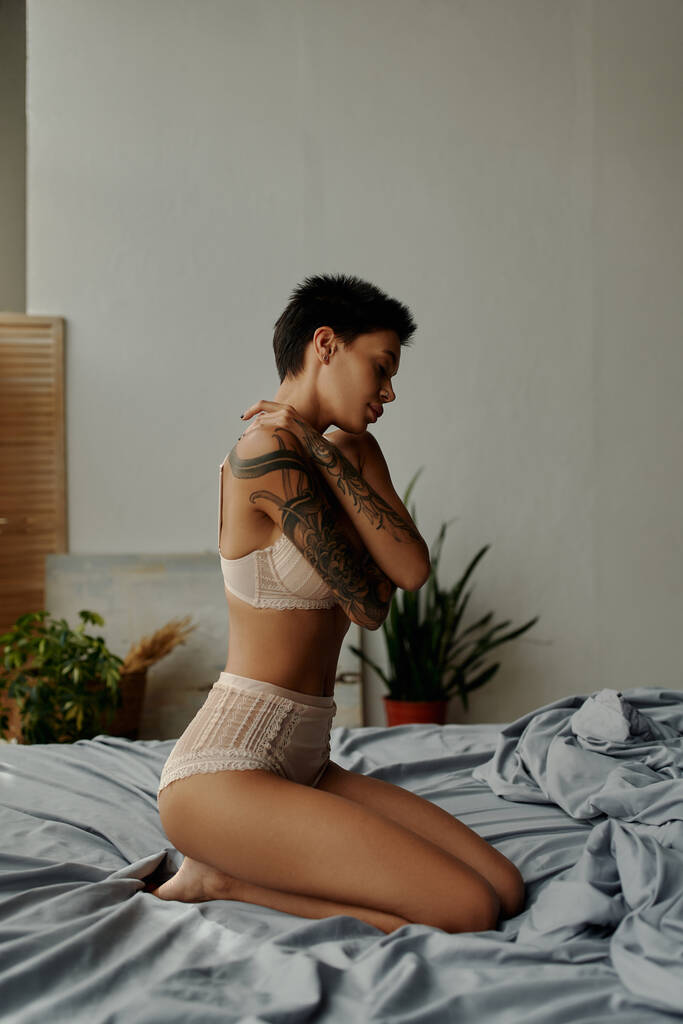 πλαϊνή άποψη της σαγηνευτικής γυναίκα τατουάζ σε εσώρουχα αγκαλιάζει τον εαυτό της, ενώ κάθεται στο κρεβάτι - Φωτογραφία, εικόνα