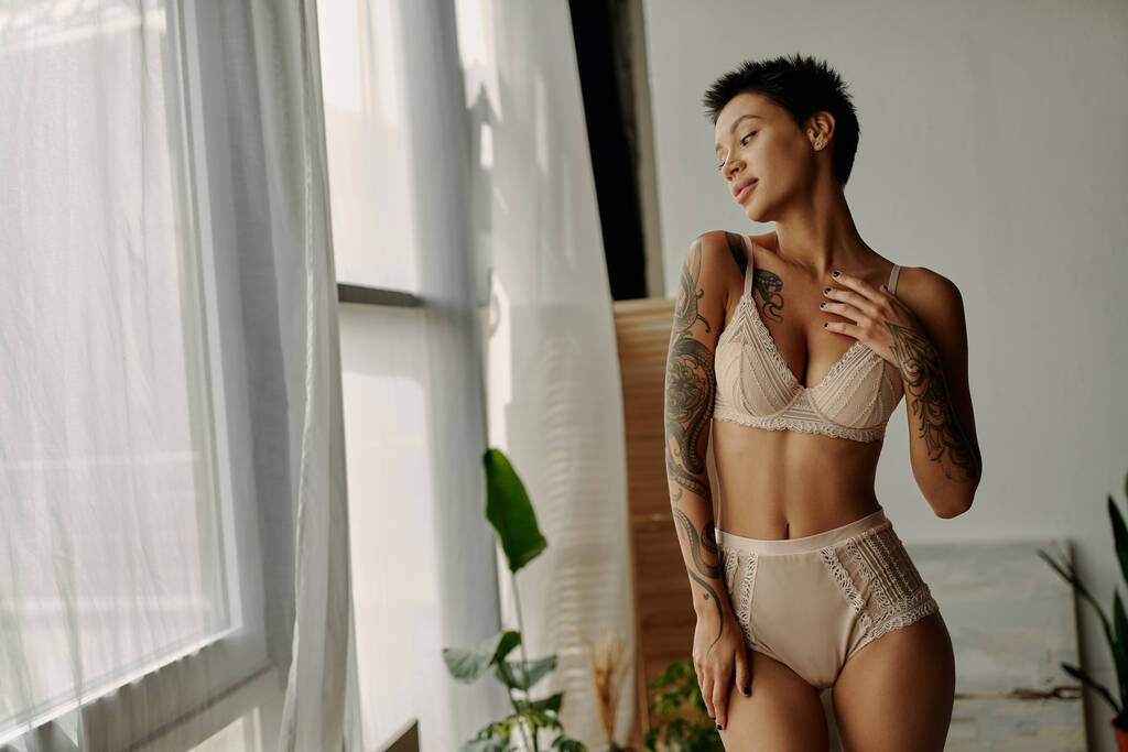 Young tattooed woman in lace lingerie looking at window at home  - Photo, Image