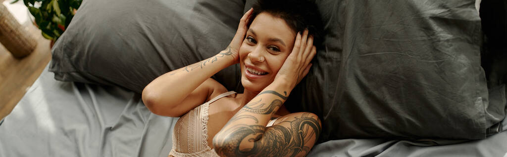 Yatakta kameraya bakan, sütyenli, dövmeli, neşeli kadının yüksek açılı görüntüsü.  - Fotoğraf, Görsel