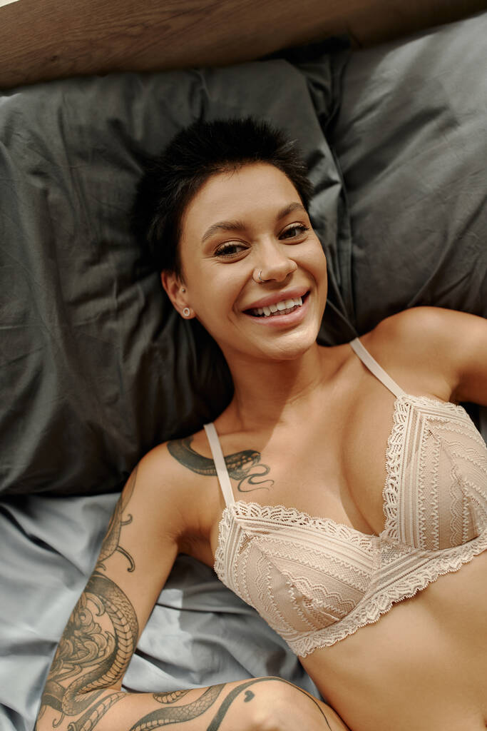 Vista dall'alto della donna tatuata positiva in reggiseno di pizzo che guarda la fotocamera mentre è sdraiato sul letto  - Foto, immagini