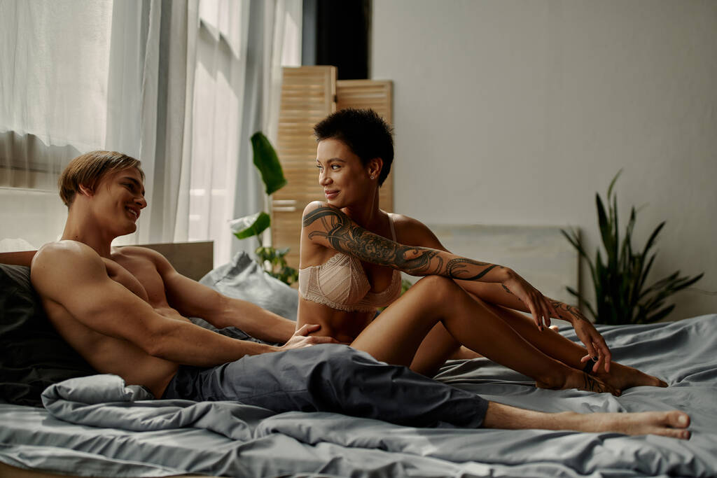 Улыбающийся мужчина в пижамных штанах смотрит на соблазнительную девушку в постели  - Фото, изображение